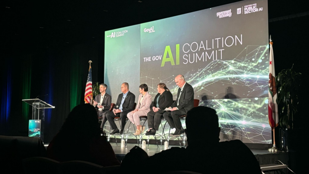 four people sitting on stage infront of a banner that reads "Gov AI Coalition Summit"